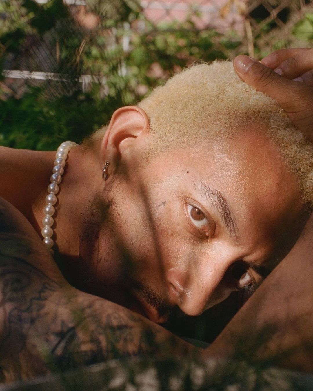 Close-up portrait of a light-skinned adult male with bleached hair and pearl necklace lying in a garden with filtered sunlight.