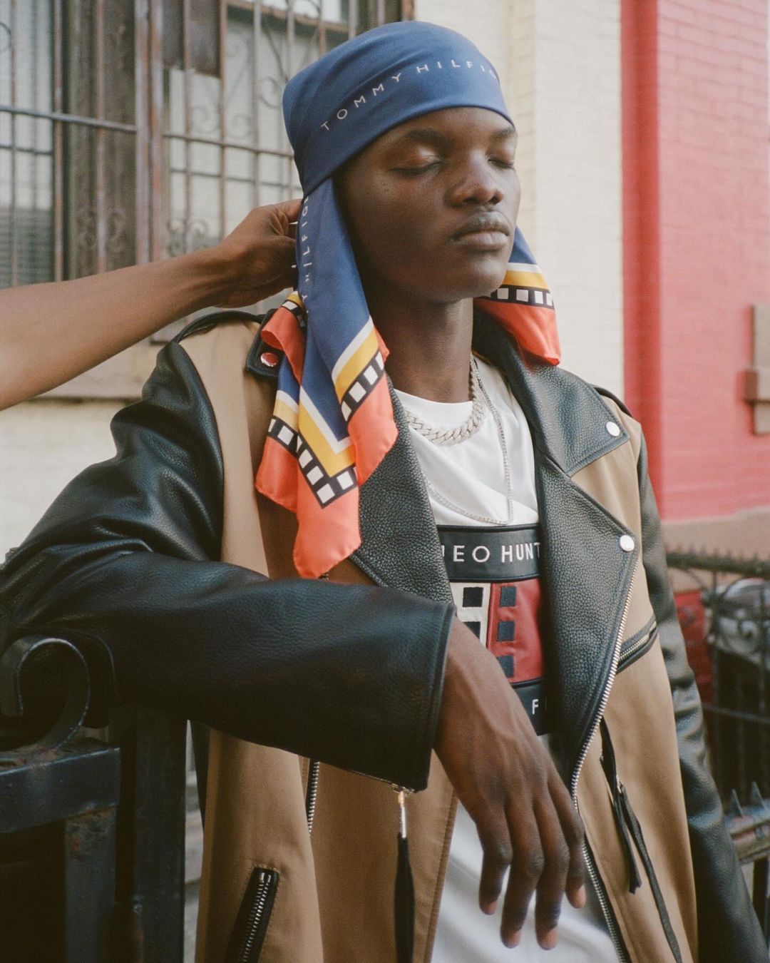 Man in headscarf and leather jacket stands outside, leaning against fence. Second person adjusts headscarf in medium shot.