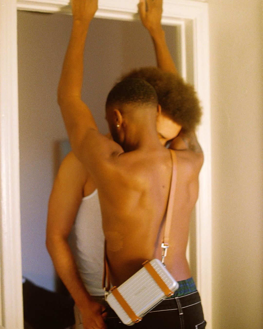 Two shirtless Black men embracing inside a doorway with arms raised, wearing a silver cross-body bag.