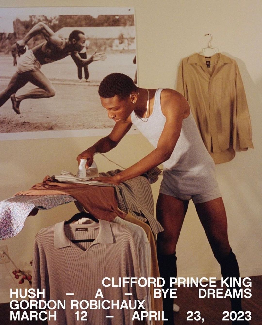An adult male irons clothes in a room with a vintage-style photo on the wall and shirts hanging. Interior shot, eye-level angle.