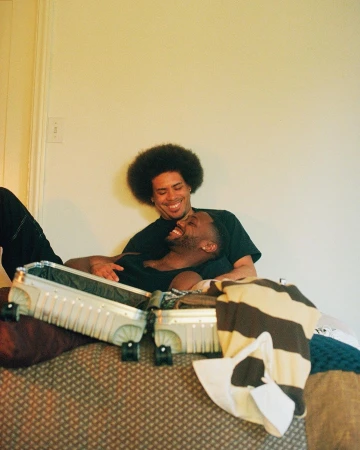 Two African-American men smiling, embracing indoors near a suitcase, creating a happy, relaxed atmosphere with casual clothing and warm lighting.
