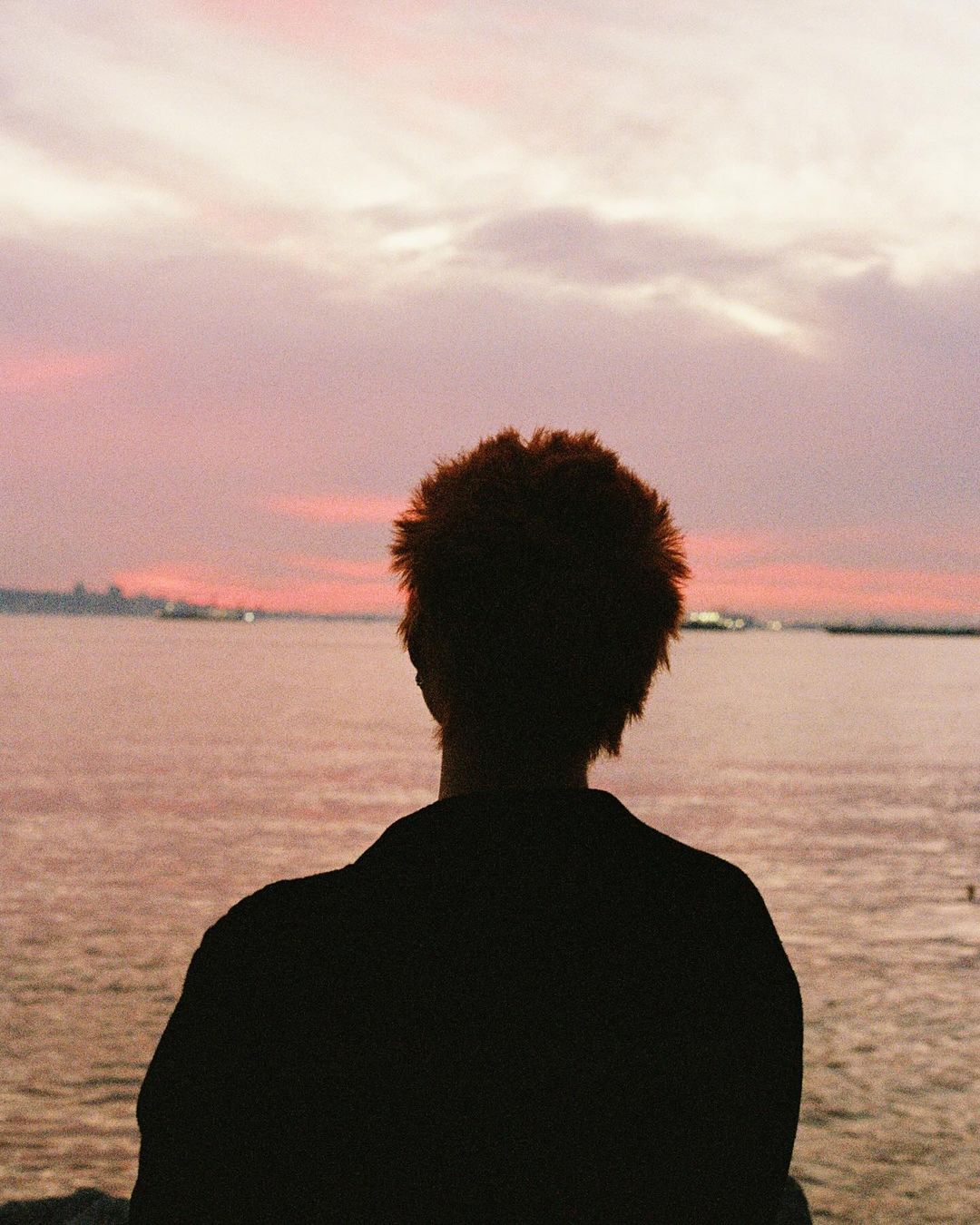 Person silhouetted against a pink and orange sunset over the water, with a distant cityscape and cloudy sky creating a scenic view.