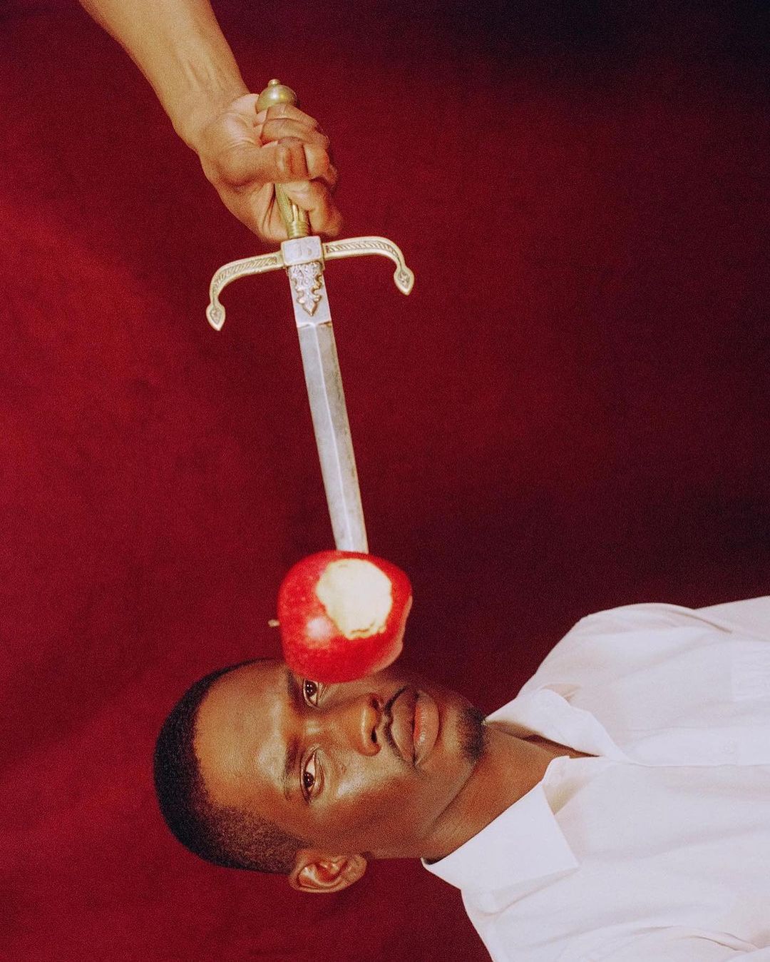 An African-American man in a white shirt poses with a sword piercing an apple balanced on his head against a red backdrop.