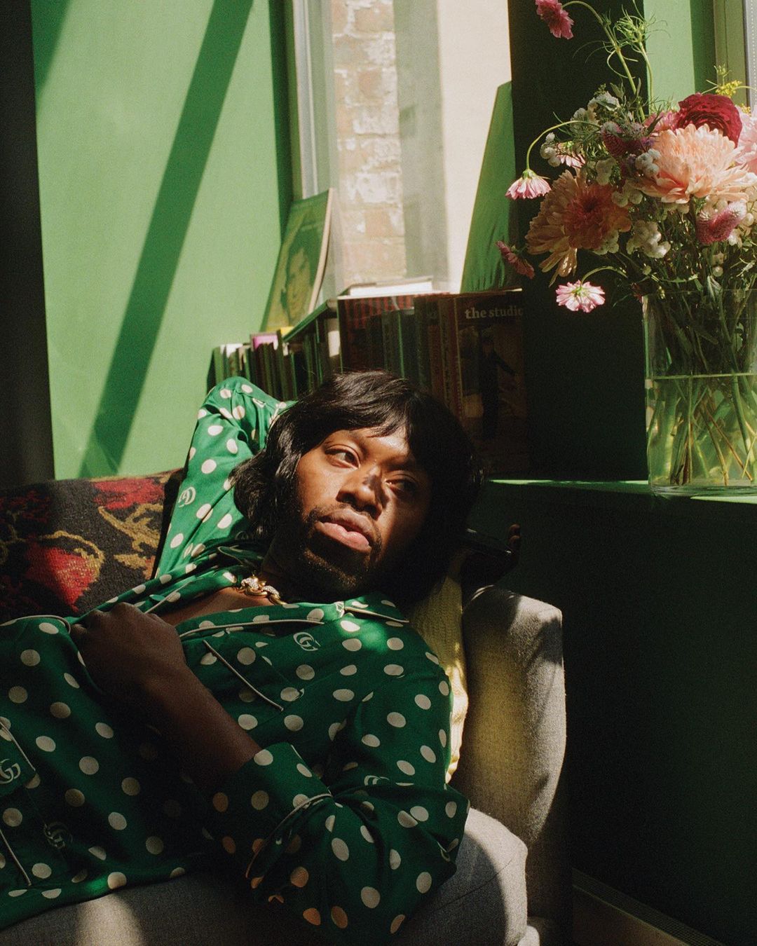 Adult lounging on a gray couch, wearing a green and white polka dot Gucci pajama set, with flowers and books in the background.