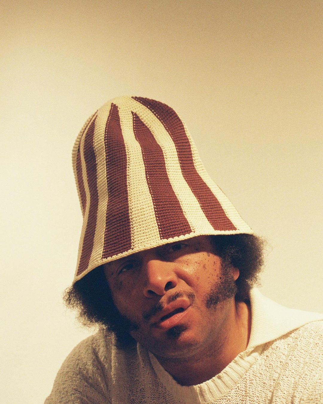 Adult male poses in a cream-colored knitted sweater and a striped crochet bucket hat in a studio with diffused lighting.