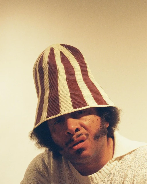Adult male poses in a cream-colored knitted sweater and a striped crochet bucket hat in a studio with diffused lighting.