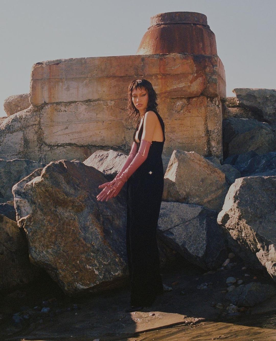 Adult woman in black dress and long pink gloves posing on the beach near large rocks in a fashion editorial style under daylight.