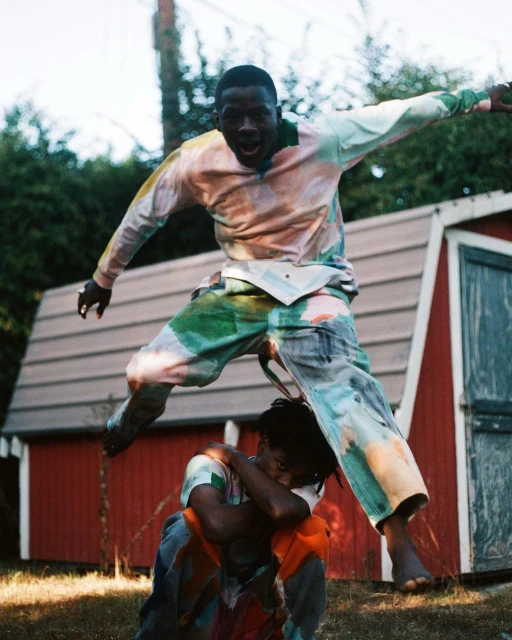 Two Black men outdoors; one jumping over the other, wearing colorful tie-dye tops and bottoms, red shed and green trees in background.