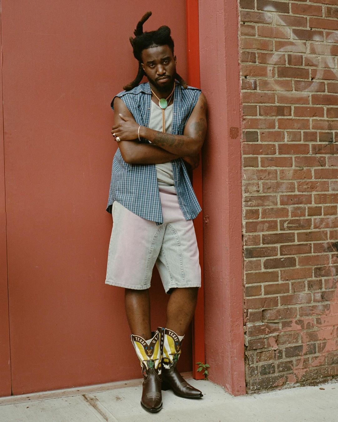 A stylish African-American man leans against a brick wall outdoors, wearing a sleeveless shirt, shorts, and cowboy boots in full view.