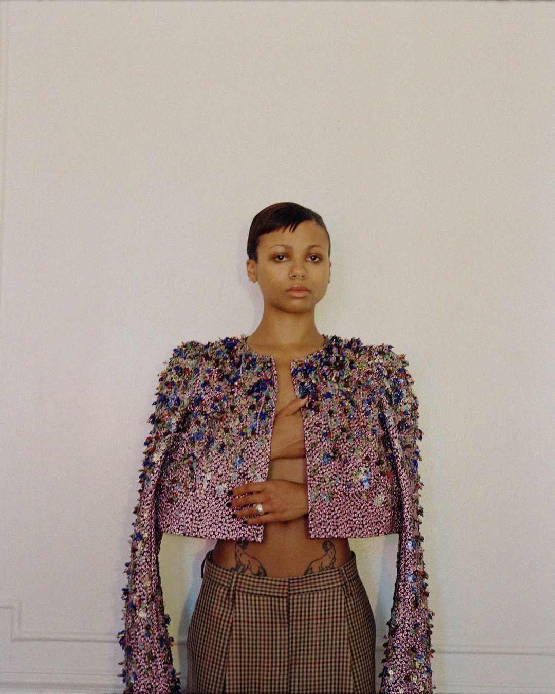 Portrait of a woman wearing a heavily embellished beaded jacket and plaid trousers against a neutral wall by Clifford Prince King