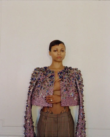 Portrait of a woman wearing a heavily embellished beaded jacket and plaid trousers against a neutral wall by Clifford Prince King
