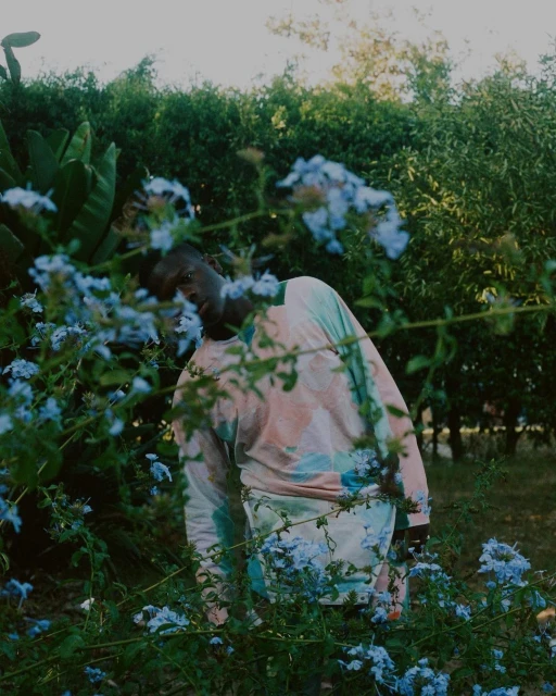 Young adult stands amid lush greenery and delicate blue flowers, wearing a colorful tie-dye shirt and shorts in a natural outdoor setting.