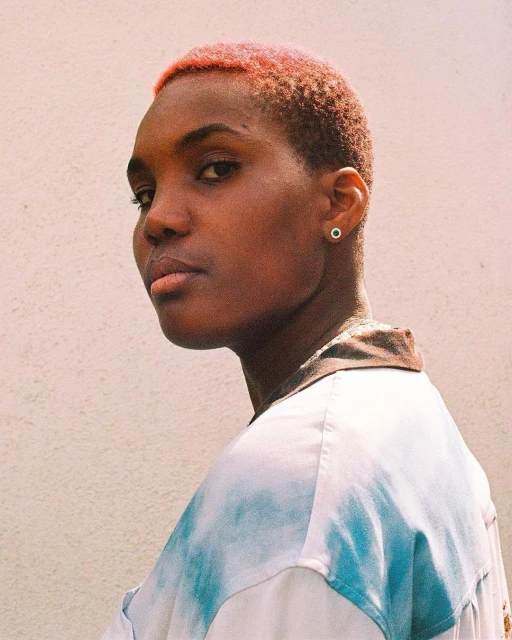 Portrait of a young adult woman with short pink hair in a white and blue tie-dye shirt, standing against a white wall.