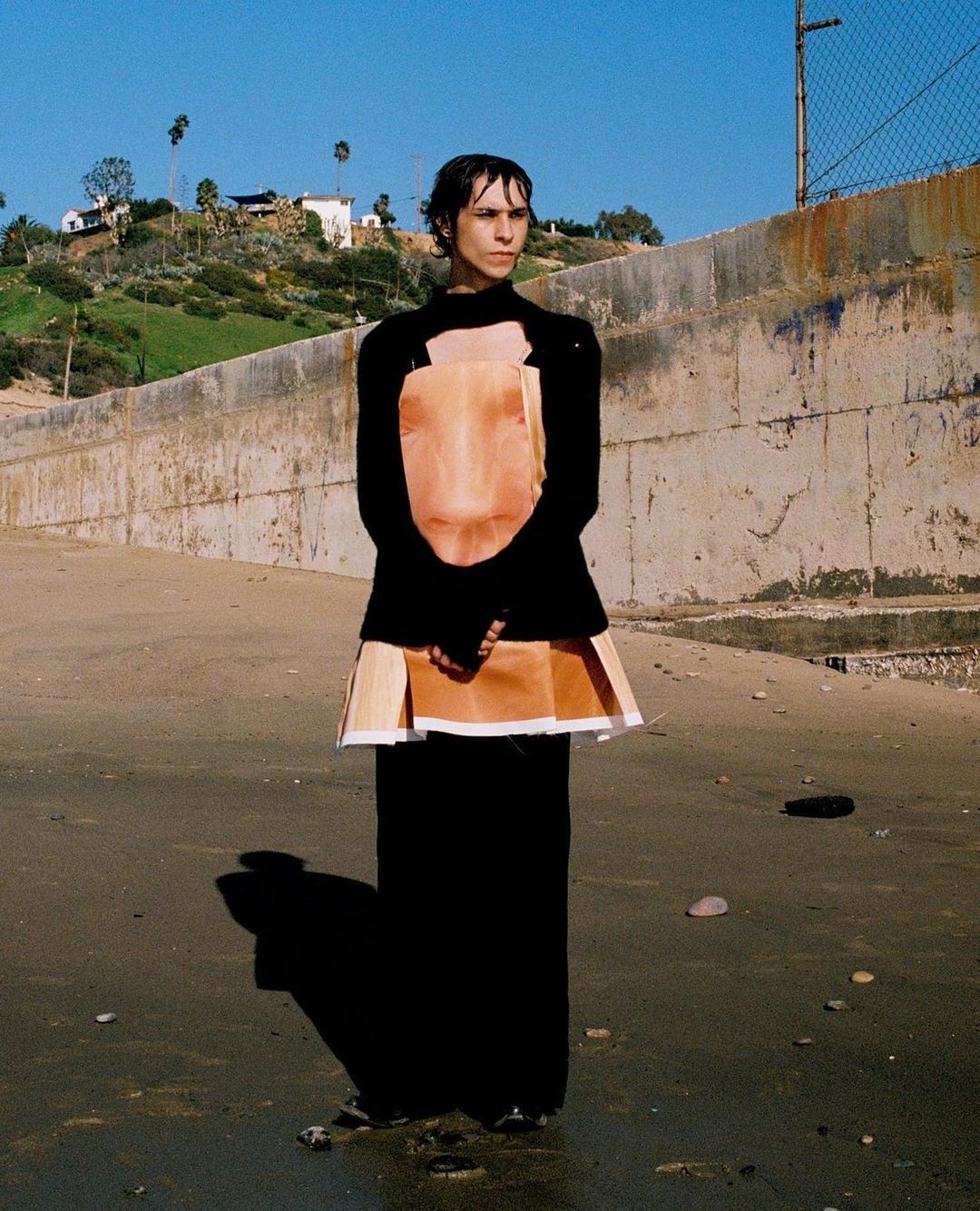 Person in a black top and long black skirt stands on a beach with a cement barrier behind them on a sunny day with a blue sky.