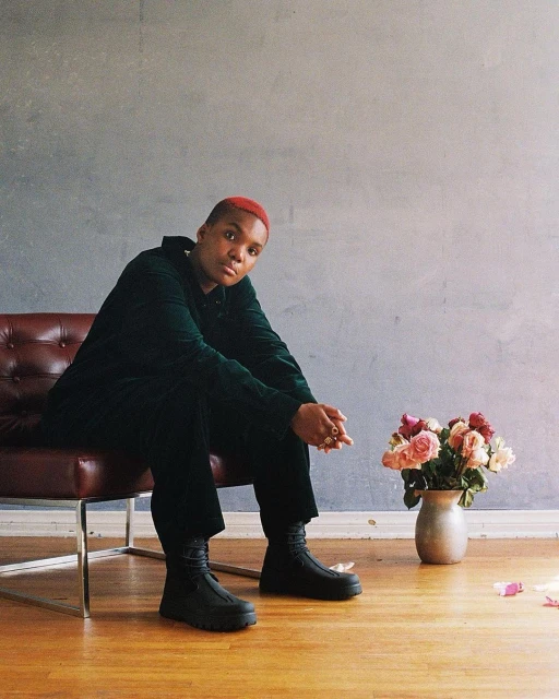 A red-haired person in a green velvet suit sits on a chair next to flowers indoors, illuminated by soft lighting against a grey wall.