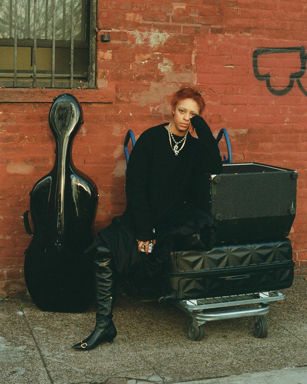 Woman with red hair wearing black sweater, dress and boots, poses with instrument case and stacked luggage against brick wall backdrop.