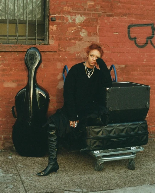 Woman with red hair wearing black sweater, dress and boots, poses with instrument case and stacked luggage against brick wall backdrop.