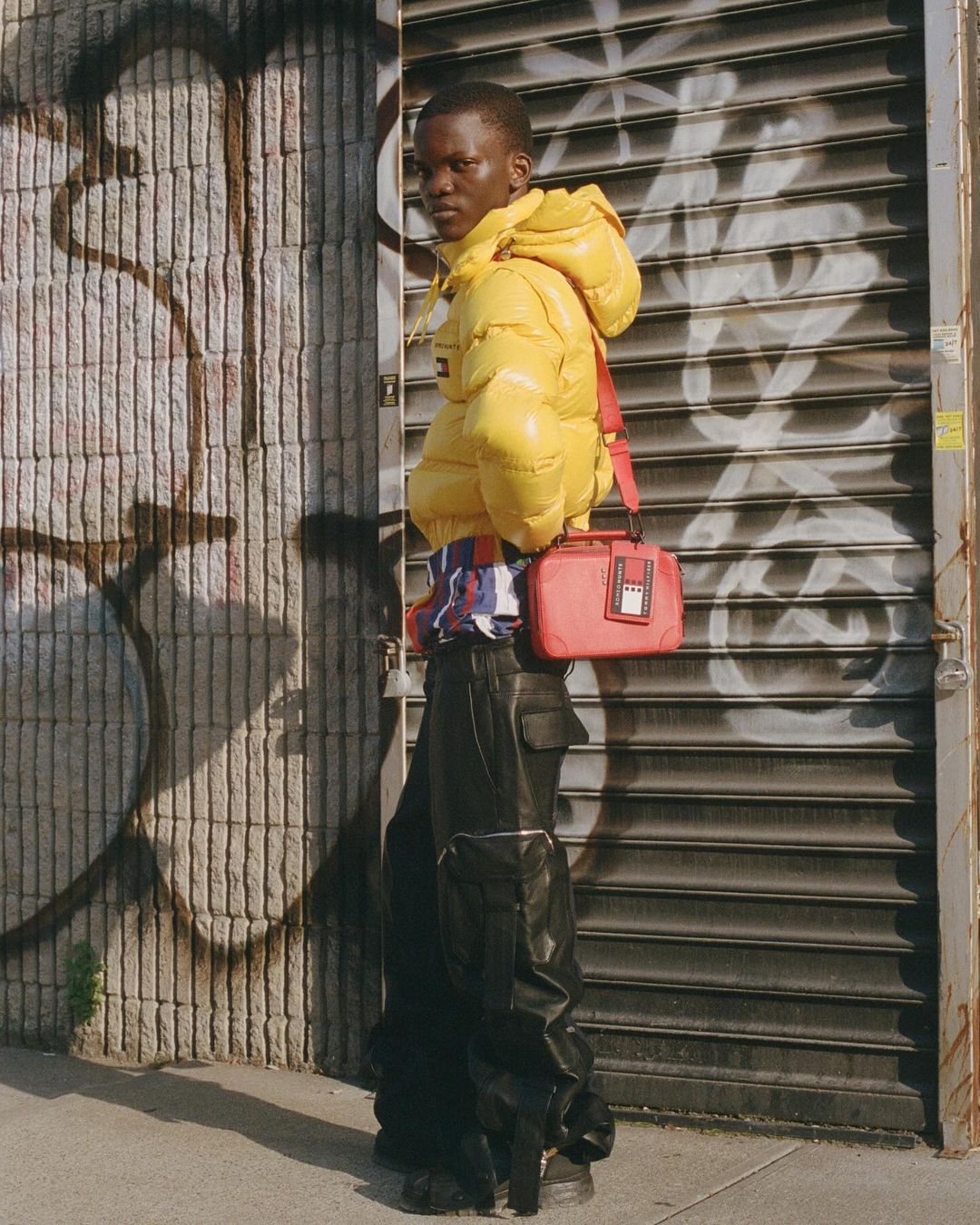 Man in yellow puffer jacket with a red bag poses outdoors against a wall with graffiti and a metal roll-up door.