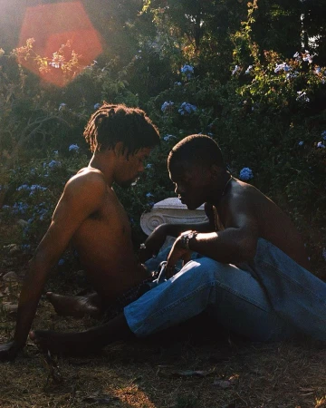 Two shirtless Black men sitting in a garden area with flowers and greenery, posing for a portrait in the sunlight, wearing blue jeans.