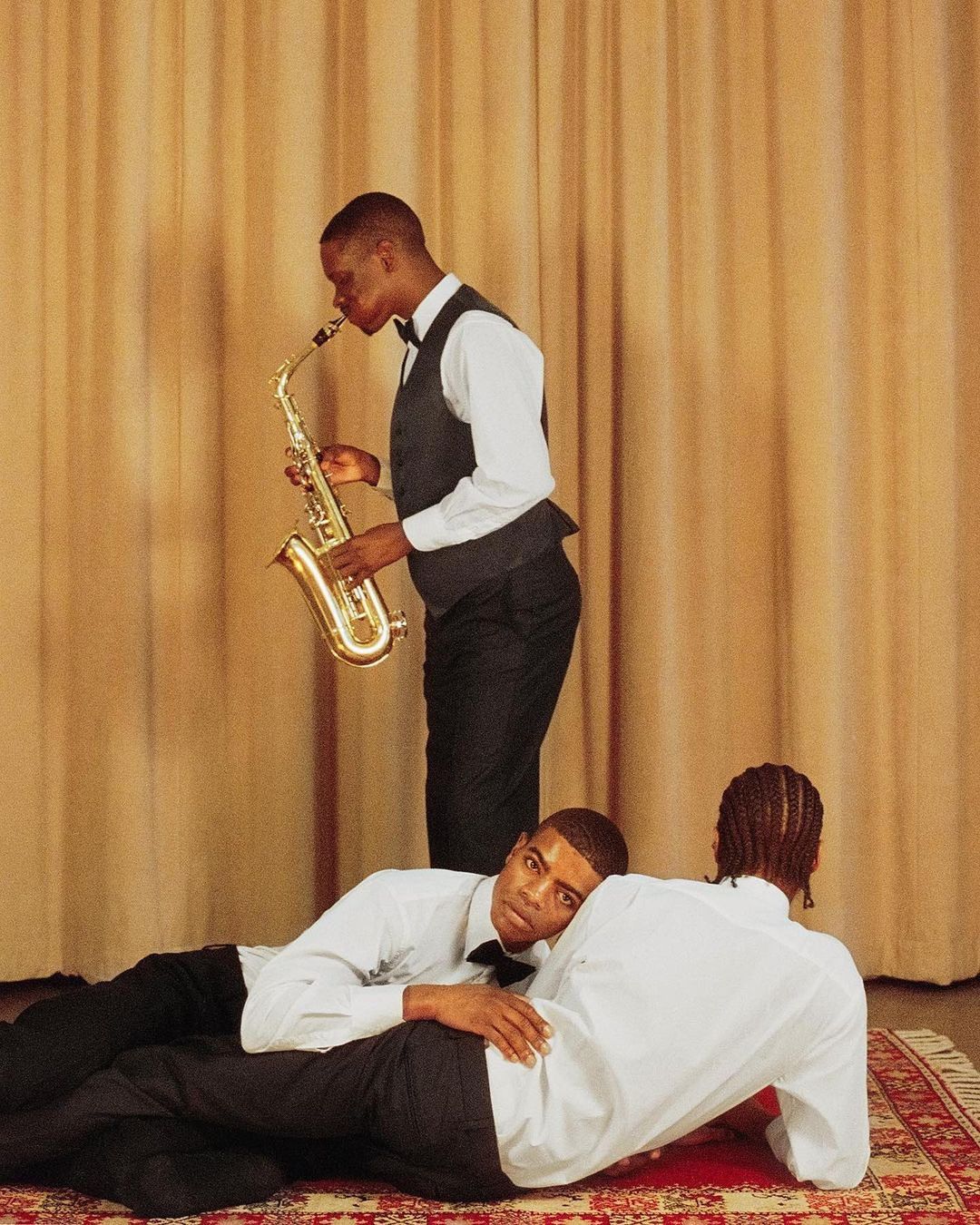 Three African American men in formal wear: one plays saxophone standing, two lying on a patterned rug against a tan curtain backdrop.
