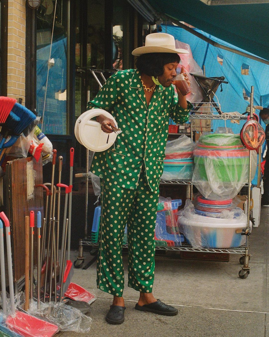 An adult wearing a green polka dot pajama set and a hat drinks from a cup outdoors near cleaning supplies and stacked bowls on a metal rack.