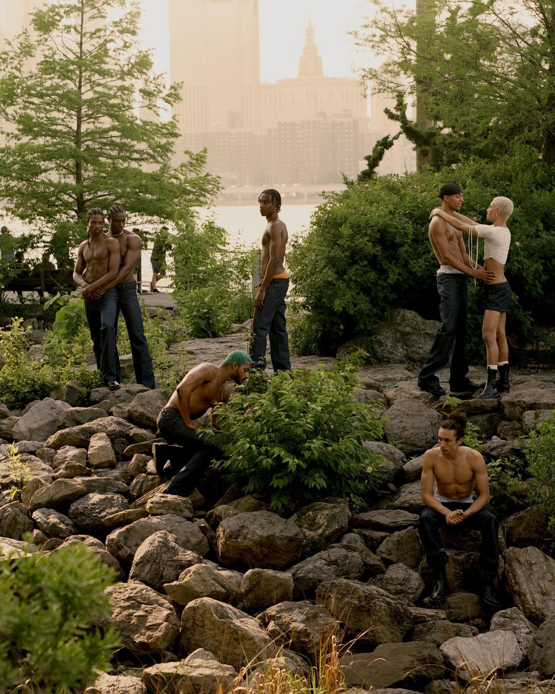 A group of shirtless men gather on a rocky riverbank with trees, with buildings visible in the hazy background during daytime.