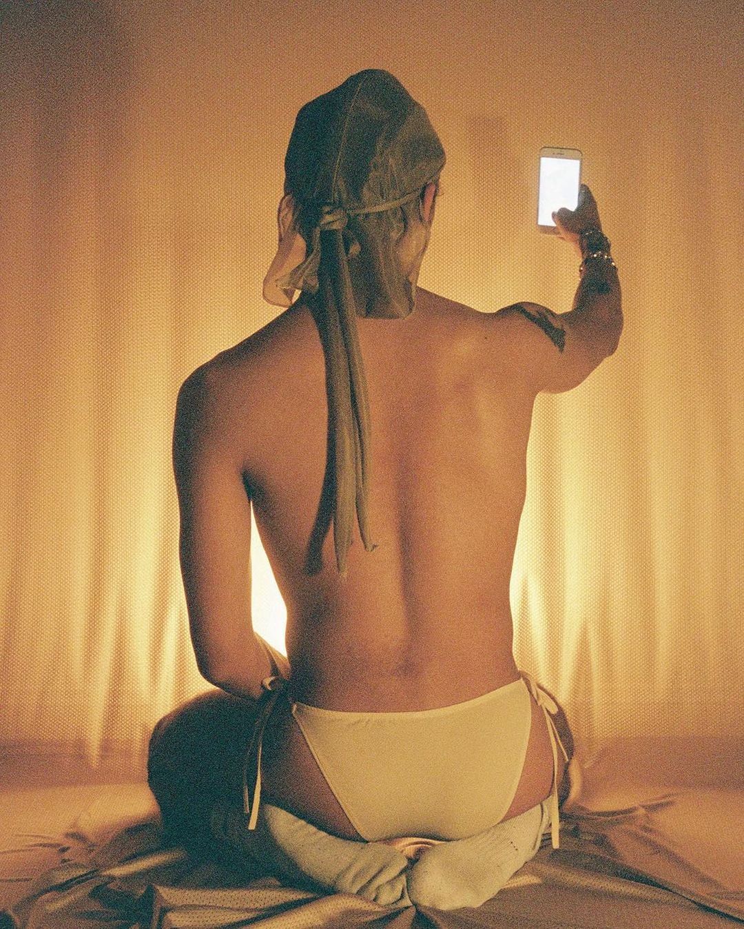 Back view of person in briefs sits cross-legged, holding up a bright cell phone, with soft golden lighting on walls and floor.