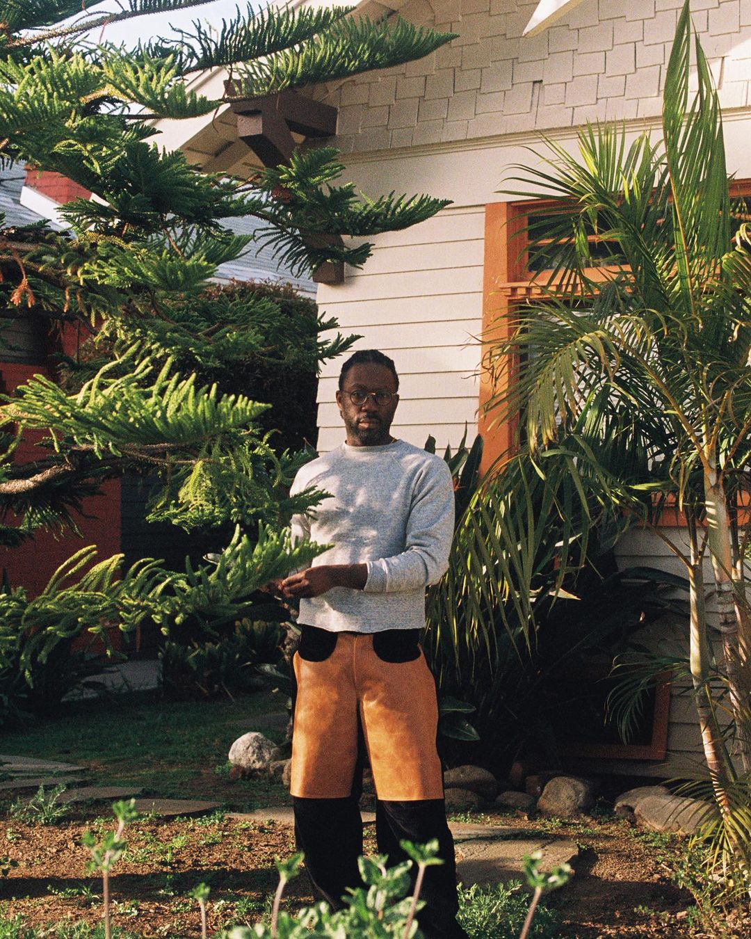 An adult male stands centered outdoors in front of a light-colored house surrounded by green plants and trees, wearing brown and black pants.