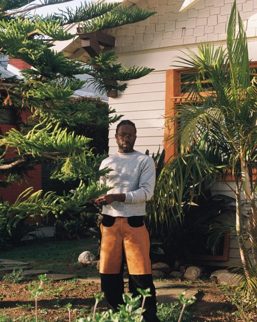An adult male stands centered outdoors in front of a light-colored house surrounded by green plants and trees, wearing brown and black pants.