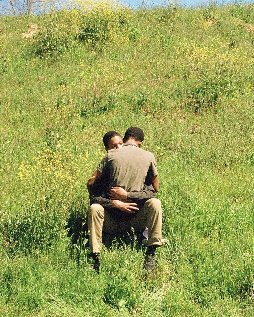 A couple embraces in a field of green grass and yellow wildflowers. The figures are seated, creating a foreground portrait in nature.