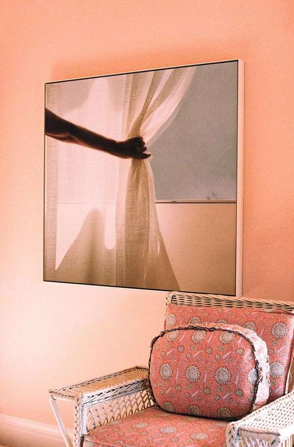 Artwork on a coral wall featuring a hand holding a curtain, above a wicker chair with patterned cushions in a peach-toned room.