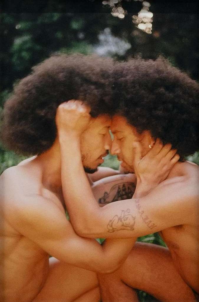 Two adult men with afros embrace outdoors with a background of foliage, showcasing a romantic and intimate connection in a natural setting.