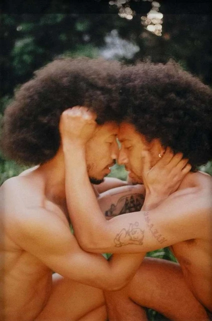 Two adult men with afros embrace outdoors with a background of foliage, showcasing a romantic and intimate connection in a natural setting.