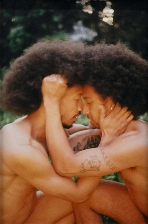 Two adult men with afros embrace outdoors with a background of foliage, showcasing a romantic and intimate connection in a natural setting.