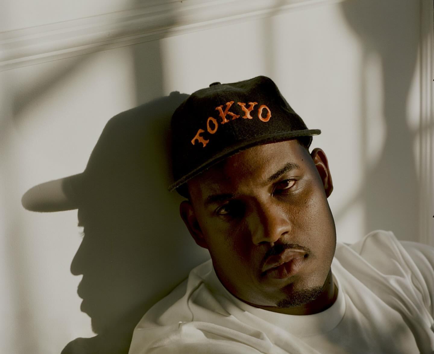 Adult male wearing a 'Tokyo' hat in a portrait featuring strong lighting and shadow play against a white wall, creating a striking visual.