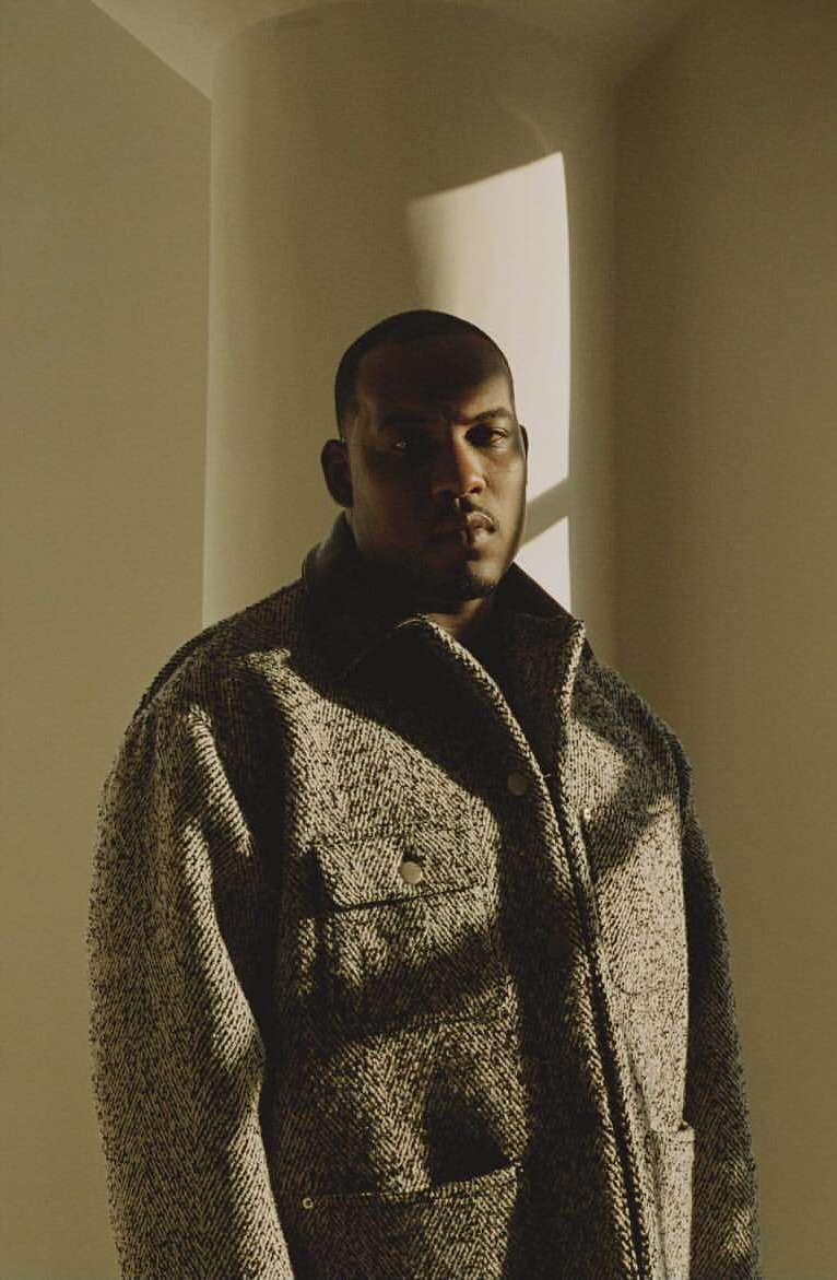 Portrait of a stylish adult male wearing a textured tweed coat inside an architectural space with diffused natural light and warm tones.