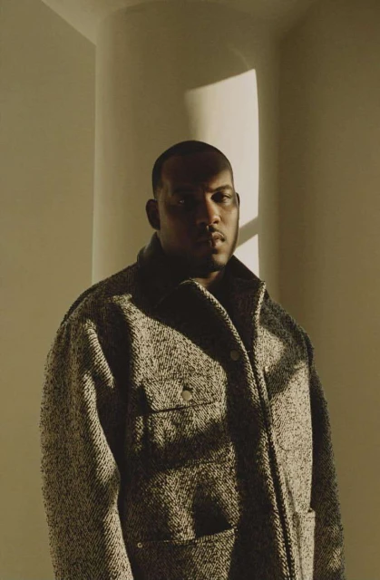 Portrait of a stylish adult male wearing a textured tweed coat inside an architectural space with diffused natural light and warm tones.