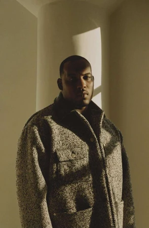 Portrait of a stylish adult male wearing a textured tweed coat inside an architectural space with diffused natural light and warm tones.