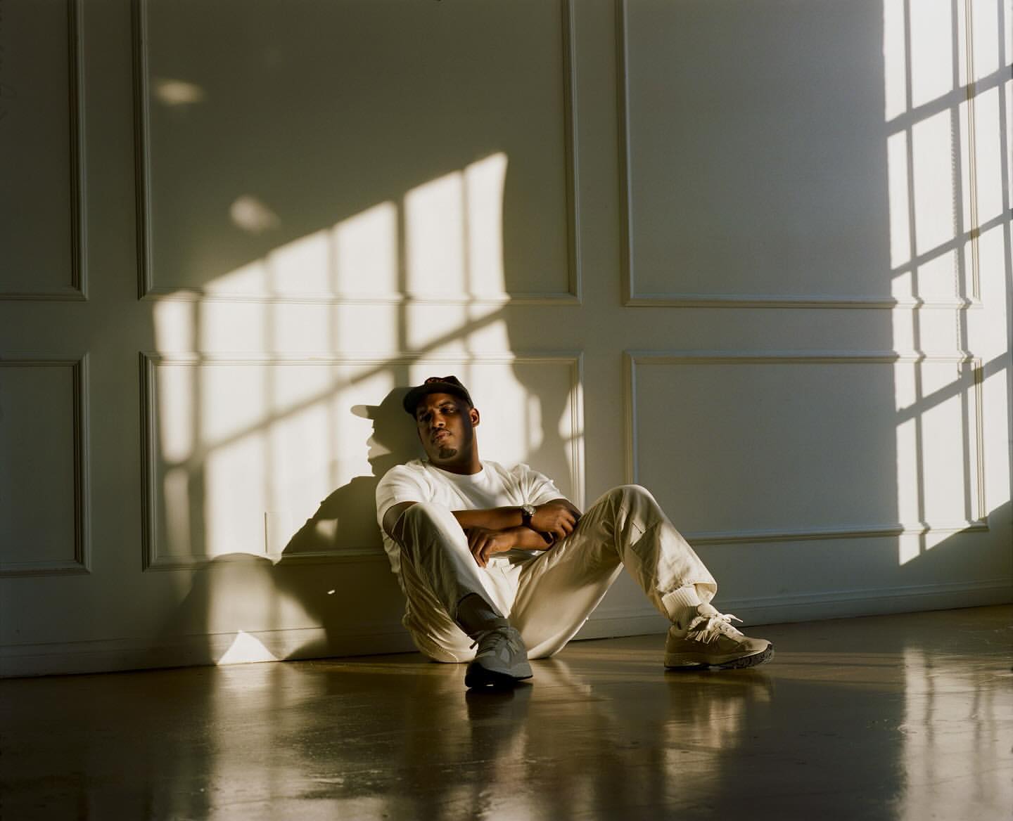 An African-American man wearing casual clothes sits on the shiny wooden floor, leaning against a panelled wall with sunlight casting shadows.