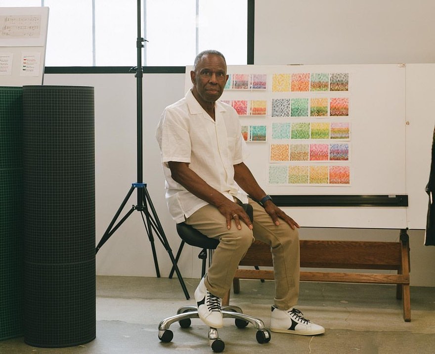 An adult man seated on a rolling chair in a studio, wearing a white shirt and khaki pants, posing in front of a whiteboard display.