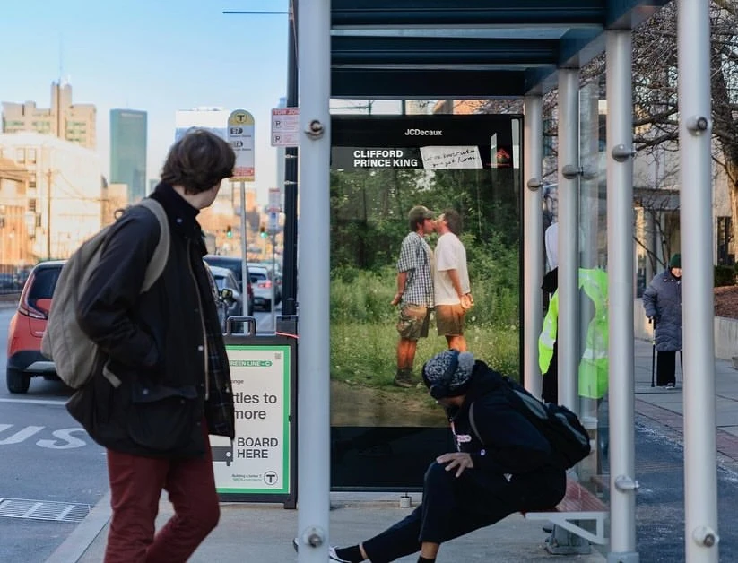 City bus stop view with people and an advertisement featuring two men embracing near green foliage on a sunny day.
