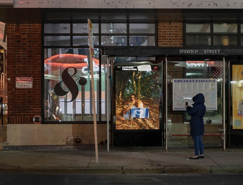 Night scene in Boston featuring a person standing at a bus stop with ads and a storefront with signage on Ipswich Street.