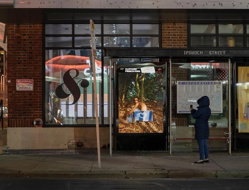 Night scene in Boston featuring a person standing at a bus stop with ads and a storefront with signage on Ipswich Street.