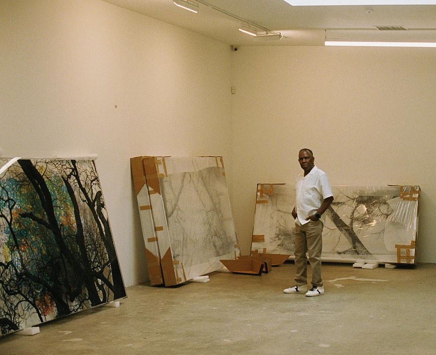 Adult male artist stands in an art gallery with large paintings, wearing a white shirt and khaki pants in a bright environment.