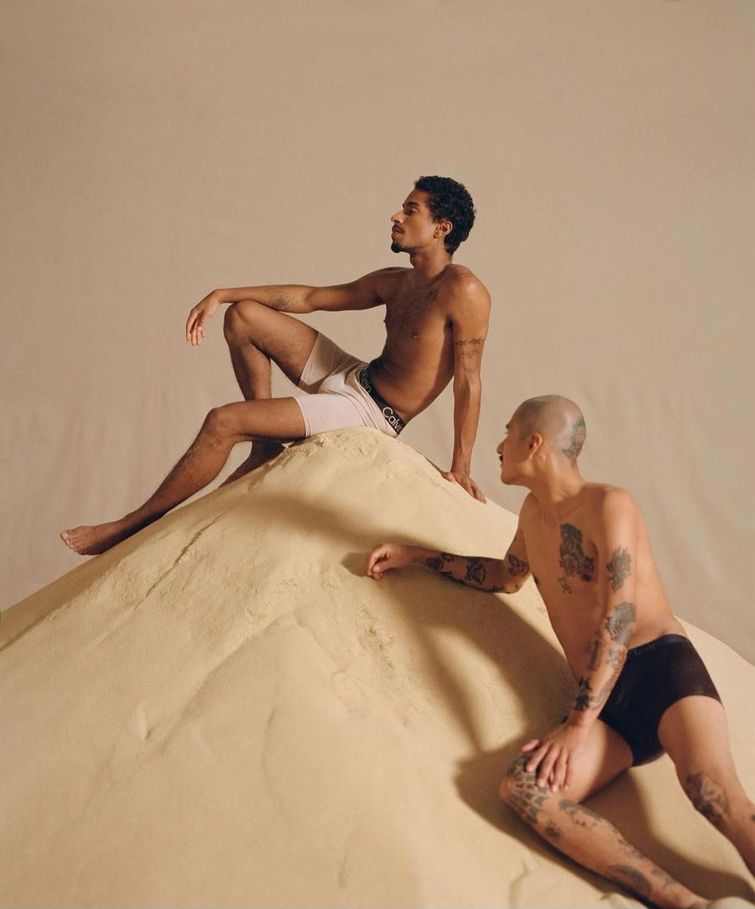Two young adult men pose on a sand mound indoors, wearing underwear in a studio setting with soft lighting and neutral tones.