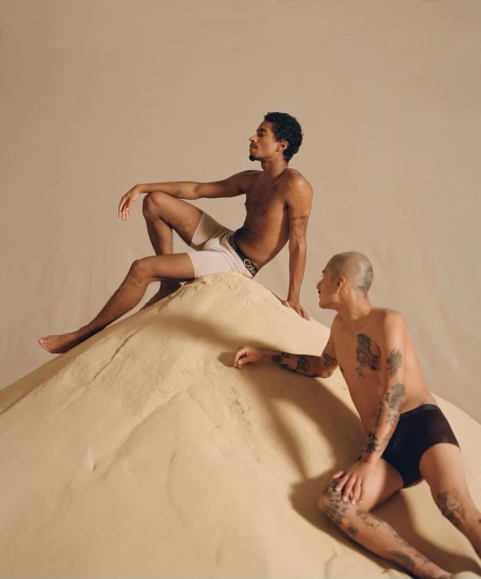 Two young adult men pose on a sand mound indoors, wearing underwear in a studio setting with soft lighting and neutral tones.