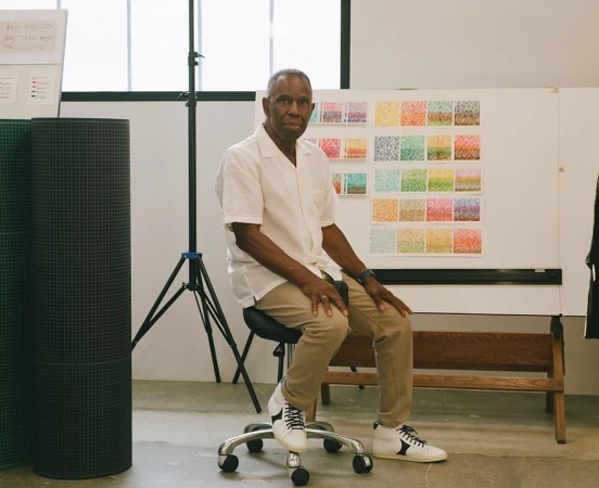 An older Black man seated on a rolling stool in a bright studio, wearing a white short-sleeve shirt and khaki trousers.