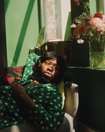 Black man in a green polka dot pajama shirt lounging on a couch next to a vase of flowers.
