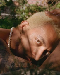 Close-up portrait of a man with blonde hair and tattoos wearing a pearl necklace outdoors in New York by Clifford Prince King.