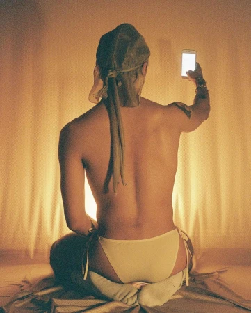 Shirtless man sitting on a bed with a head covering holding up a lit smartphone to take a photo, shot by Clifford Prince King.